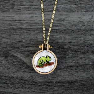 May include: A gold chain necklace with a round wooden pendant featuring a green embroidered chameleon.