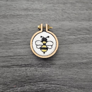 Puede incluir: Un pequeño aro de madera redondo con un centro de tela blanca que presenta una abeja bordada en negro y amarillo.
