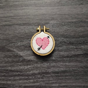 Puede incluir: Un pequeño corazón rosa con una flecha que lo atraviesa está bordado en un círculo de tela blanca, enmarcado en un aro de madera.
