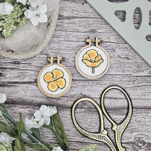 Könnte beinhalten: Zwei kleine runde Stickrahmen aus Holz mit Kreuzstichmotiven gelber Blumen auf weißem Stoff. Jeder Rahmen verfügt über eine Metallschraube zum Festziehen an der Oberseite. Die Stickereien werden zusammen mit einer Metallschere präsentiert.