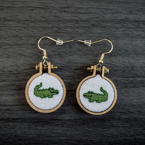 May include: A pair of earrings featuring a green alligator cross-stitch design on a white background, framed in a round wooden hoop.