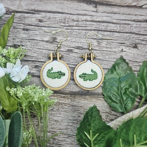 Könnte beinhalten: Ein Paar Ohrhänger mit kleinen, runden Stickrahmen aus Holz. Jeder Rahmen enthält weißen Stoff mit einem kleinen, grün im Kreuzstich gestickten Alligator-Motiv. Die Ohrringe sind mit goldfarbenen Metallhaken versehen.