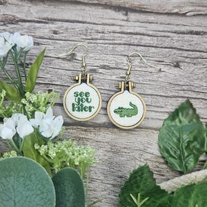May include: A pair of dangle earrings featuring miniature circular wooden embroidery hoops. One hoop contains white fabric with the green stitched text see you later, while the other features a green stitched alligator. Both have gold-tone hooks.