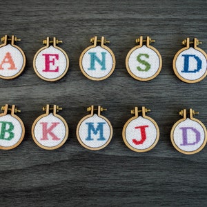 May include: A collection of ten small, round embroidery hoops, each featuring a different letter of the alphabet. The letters are stitched in various colors, including red, green, blue, and pink, against a white background. The hoops are made of wood.