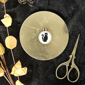 Könnte beinhalten: Ein schwarzes Katzen-Kreuzstich-Design auf einem runden weißen Hintergrund mit einem goldenen Metallrahmen. Das Design befindet sich auf einem kleinen, runden, goldfarbenen Teller. Eine goldfarbene Schere auf schwarzem Hintergrund.