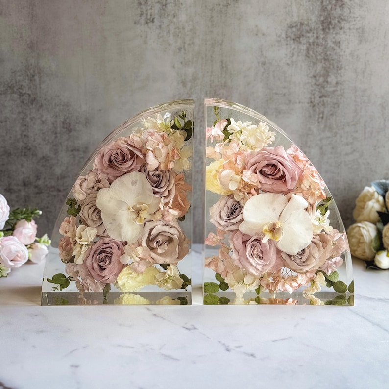 May include: Two clear, semi-circular bookends filled with dried flowers. The floral arrangements include roses, orchids, and hydrangeas in shades of pink, cream, and peach. The bookends are displayed on a white marble surface.