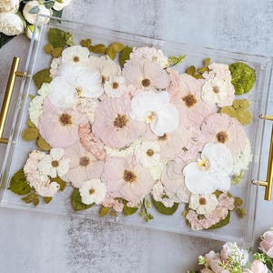 May include: A clear acrylic tray with gold handles, filled with an arrangement of dried flowers in shades of pink, white, and cream. Green leaves are interspersed throughout the floral display, creating a natural aesthetic.
