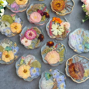 May include: A collection of clear resin coasters with dried flowers embedded within. Each coaster features a unique arrangement of colorful blooms, including yellow, pink, and orange flowers, with gold leaf accents. The coasters have a wavy edge.