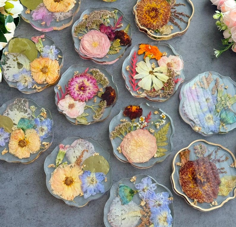 May include: A collection of clear resin coasters with dried flowers embedded within. Each coaster features a unique arrangement of colorful blooms, including yellow, pink, and orange flowers, with gold leaf accents. The coasters have a wavy edge.