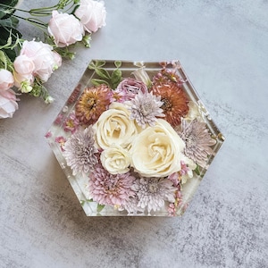 May include: A clear, hexagonal resin coaster with a floral arrangement. The coaster features cream-colored roses, lavender chrysanthemums, and orange dahlias. The flowers are encased in clear resin, creating a decorative and functional item.