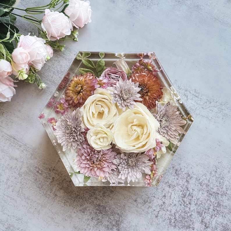 May include: A clear, hexagonal resin coaster with a floral arrangement. The coaster features cream-colored roses, lavender chrysanthemums, and orange dahlias. The flowers are encased in clear resin, creating a decorative and functional item.