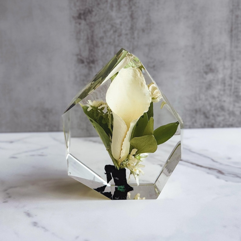 May include: A clear, geometric resin block encasing a white calla lily and greenery. The flower is the focal point, with its creamy petals and green leaves preserved within the transparent resin. Small white flowers add detail.