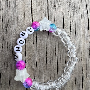 May include: A clear plastic bracelet with a star charm and letter beads spelling "ROHA 7". The beads are white with black lettering. The star charm is white with a glittery blue and pink centre.
