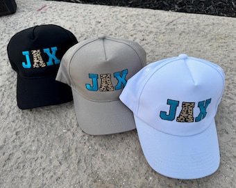 May include: Three baseball caps in black, gray, and white. Each cap features the word "JAX" embroidered in teal with a leopard print fill. The caps are displayed on a concrete surface.
