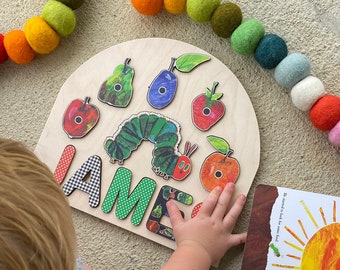 Puede incluir: Un rompecabezas de madera con la oruga muy hambrienta con una oruga colorida y piezas de fruta. El rompecabezas tiene forma de arco y tiene el nombre "JAME" escrito en letras de colores. También se muestra un libro con los mismos personajes.