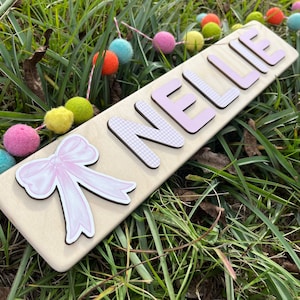 May include: A wooden sign with the name "NELLIE" spelled out in pink letters with a gingham pattern. A pink bow is on the left side of the sign.