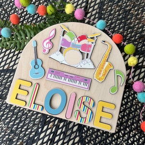 May include: Wooden name puzzle with the name Eloise. The puzzle features a colorful musical theme with a guitar, keyboard, drums, saxophone, and a musical note.