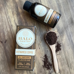 May include: A brown bottle with a black cap and a white label that says "HALO PANTRY™ SMALL BATCH VIETNAMESE COFFEE ALL NATURAL FLAVORING ZERO CALORIES | ZERO SUGARS 2 FL OZ (60ml)". The bottle is sitting on a wooden surface. There is a wooden spoon with coffee grounds in it and coffee grounds scattered around the spoon.