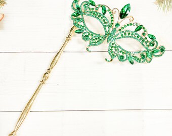 May include: A gold masquerade mask with a butterfly design. The mask is decorated with green rhinestones and is attached to a gold stick.