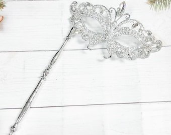 May include: A silver masquerade mask with a butterfly design, adorned with rhinestones. The mask is attached to a long silver stick.