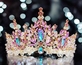 May include: A gold-coloured crown adorned with pink, purple, and teal gemstones. The crown has a symmetrical design with leaf-like details and clear crystal embellishments. The gemstones are teardrop and oval shaped.