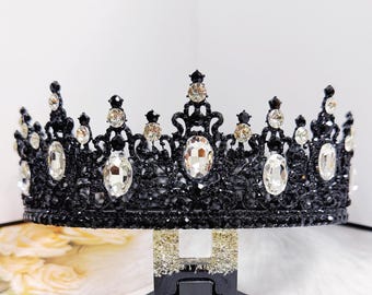 May include: A black crown with clear, oval-shaped and round gemstones. The crown is ornate with a filigree design and is displayed on a black stand. The gemstones are set in a symmetrical pattern, adding sparkle and elegance.