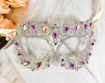 May include: A sparkly masquerade mask with iridescent rhinestones and a delicate, intricate design. The mask is made of metal and has a white background.