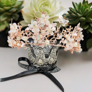 May include: A rose gold tiara with white flower and butterfly accents, and faux pearls. The crown sits atop a silver decorative crown-shaped holder with a black ribbon tied in a bow.