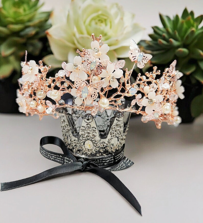 May include: A rose gold tiara with white flower and butterfly accents, and faux pearls. The crown sits atop a silver decorative crown-shaped holder with a black ribbon tied in a bow.