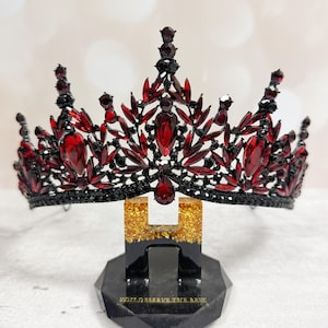 May include: A black and red jeweled crown with a decorative design. The crown is displayed on a black stand with gold accents and the text "YOU DESERVE THE BEST". The crown features large red gemstones.