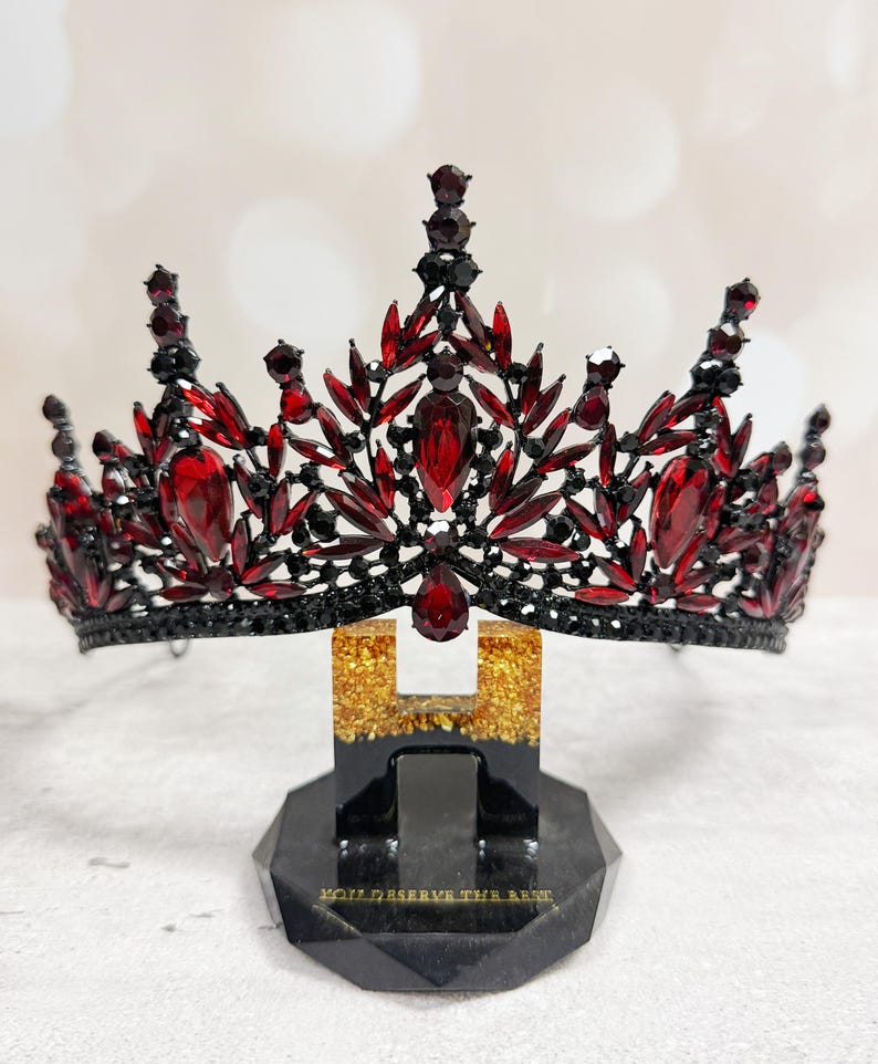 May include: A black and red jeweled crown with a decorative design. The crown is displayed on a black stand with gold accents and the text "YOU DESERVE THE BEST". The crown features large red gemstones.