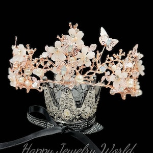 May include: A rose gold crown with white floral accents, pearls, and butterfly details. The crown sits atop a decorative silver crown-shaped holder with a black ribbon. The text "Happy Jewelry World" is visible.