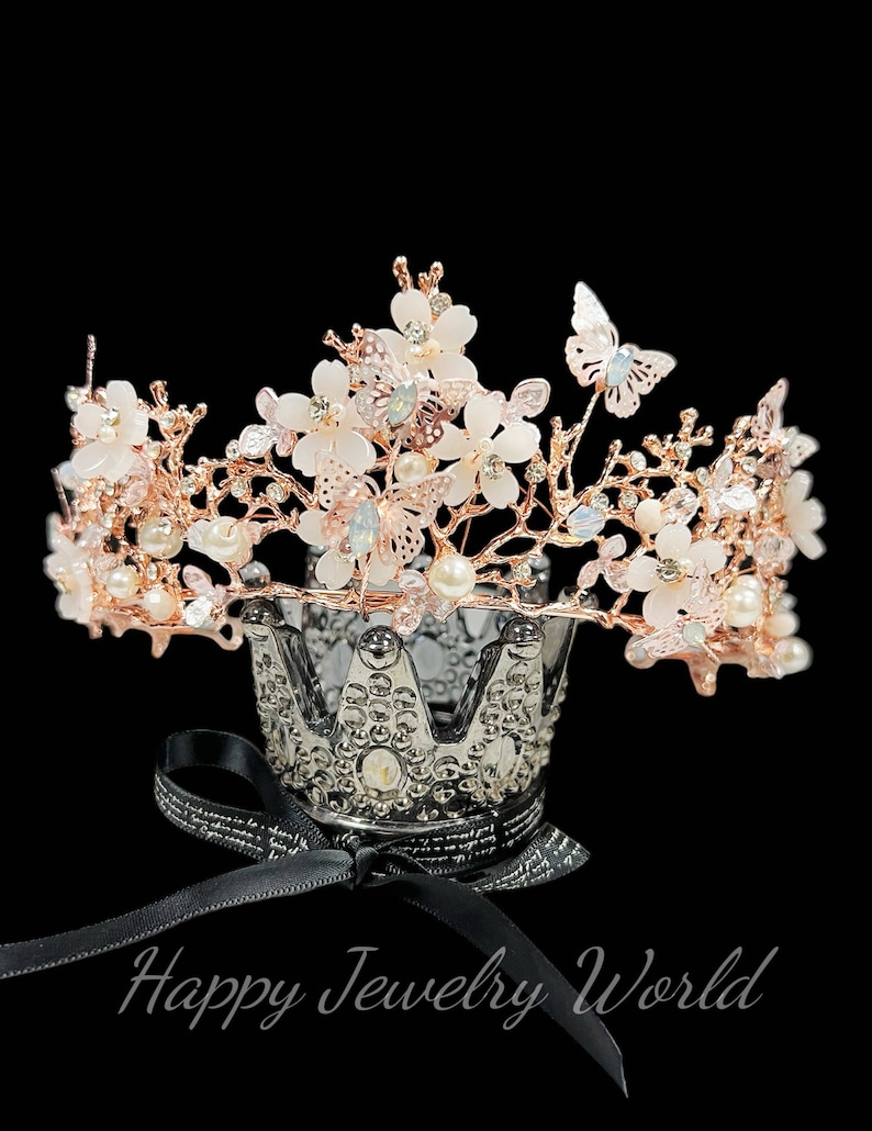 May include: A rose gold crown with white floral accents, pearls, and butterfly details. The crown sits atop a decorative silver crown-shaped holder with a black ribbon. The text "Happy Jewelry World" is visible.