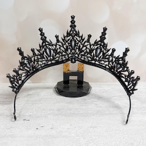 May include: A black tiara with a decorative filigree design and clear gemstones. The crown is displayed on a black stand with gold glitter accents. The tiara has a curved band and pointed accents.