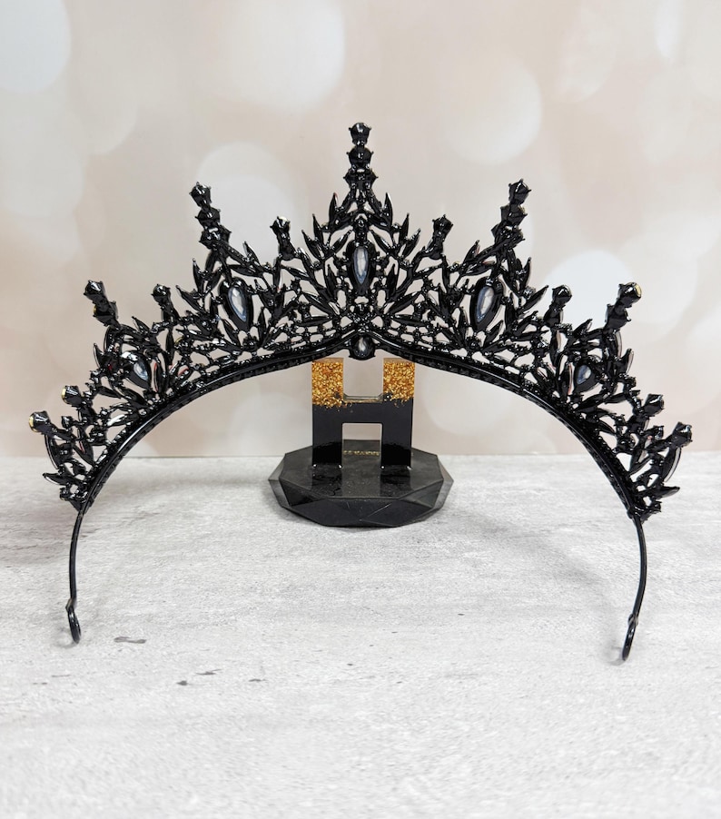 May include: A black tiara with a decorative filigree design and clear gemstones. The crown is displayed on a black stand with gold glitter accents. The tiara has a curved band and pointed accents.