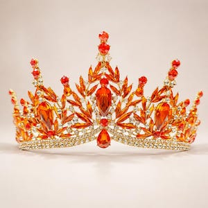 May include: A gold-toned crown featuring a base of clear rhinestones and an intricate arrangement of orange and red teardrop-shaped crystals. The design includes multiple vertical points topped with small red crystals and leaf-like accents.