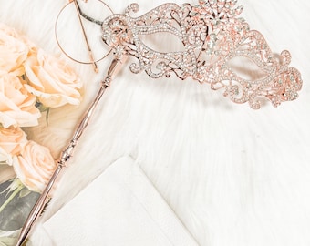 May include: A rose gold masquerade mask with intricate detailing and rhinestones. The mask is attached to a long, rose gold stick.