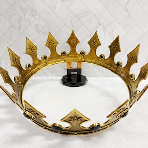 May include: A gold-colored crown with a detailed design. The crown features pointed arches and is adorned with black accents. The crown is displayed on a white surface with a patterned background.