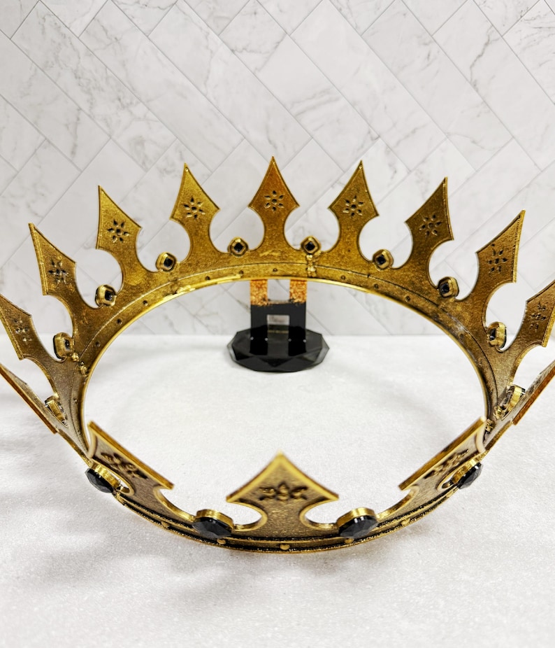 May include: A gold-colored crown with a detailed design. The crown features pointed arches and is adorned with black accents. The crown is displayed on a white surface with a patterned background.