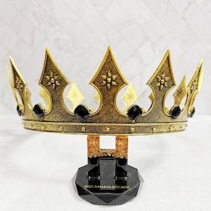 May include: A gold-colored crown with black accents and a decorative base. The crown features pointed arches, floral details, and black gems. The base is black with the text "YOU DESERVE THE BEST".