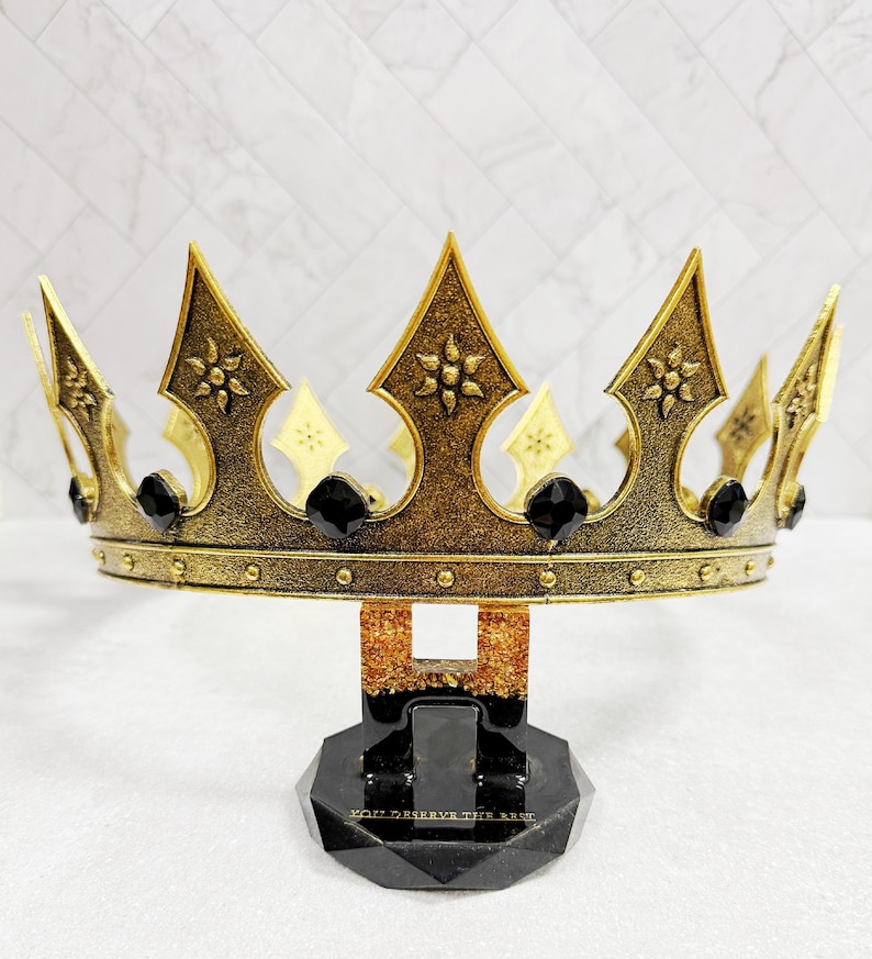 May include: A gold-colored crown with black accents and a decorative base. The crown features pointed arches, floral details, and black gems. The base is black with the text "YOU DESERVE THE BEST".