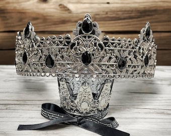 May include: A silver crown with black gemstones and intricate detailing. The crown is on a wooden surface with a black ribbon.
