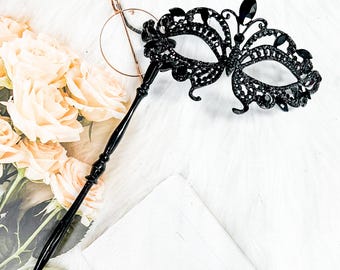 May include: A black masquerade mask with a butterfly design and rhinestones. The mask is attached to a black stick.