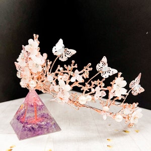 May include: A rose gold bridal crown featuring delicate white flowers, faux pearls, and butterfly accents. The crown is displayed on a purple pyramid-shaped object. The design is elegant and suitable for weddings or special occasions.