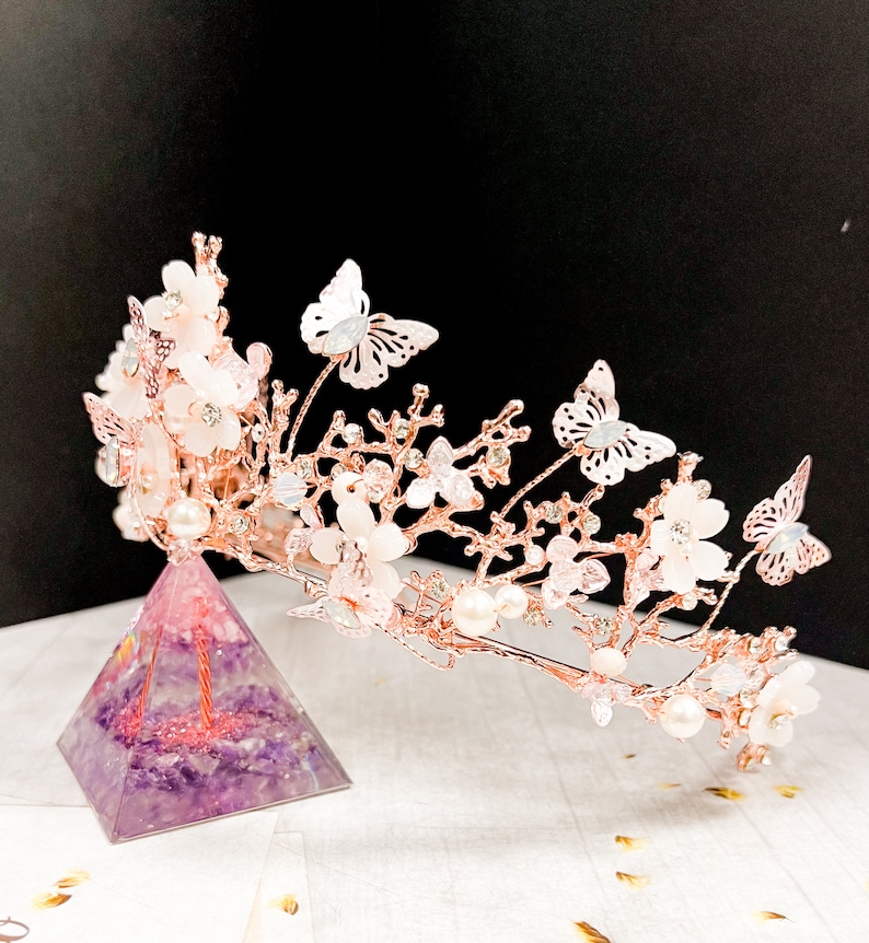 May include: A rose gold bridal crown featuring delicate white flowers, faux pearls, and butterfly accents. The crown is displayed on a purple pyramid-shaped object. The design is elegant and suitable for weddings or special occasions.