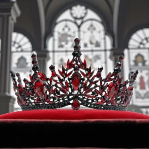 May include: A regal crown with a black metal frame, adorned with vibrant red gemstones. The crown is set on a plush red velvet cushion, showcasing its intricate design and craftsmanship. The background features a blurred architectural setting.