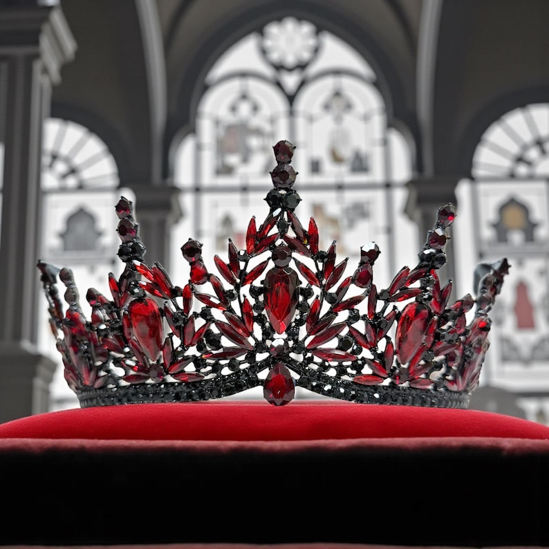 May include: A regal crown with a black metal frame, adorned with vibrant red gemstones. The crown is set on a plush red velvet cushion, showcasing its intricate design and craftsmanship. The background features a blurred architectural setting.