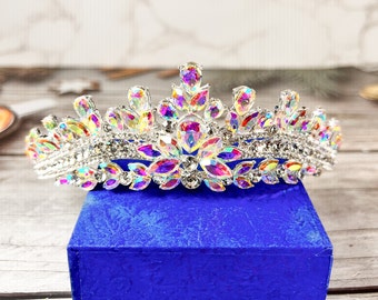 May include: A silver tiara with iridescent rhinestones and a blue velvet box. The tiara has a pointed crown and is decorated with a variety of crystal shapes.