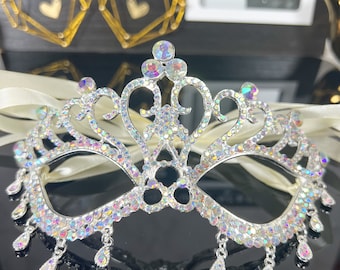 May include: A silver masquerade mask with iridescent rhinestones and a delicate, ornate design. The mask has a crown-like top and dangling teardrop-shaped accents.
