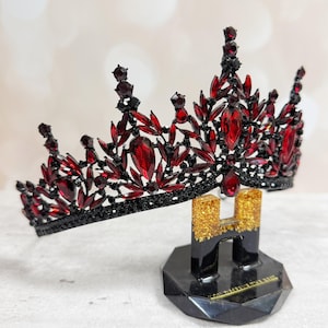 May include: A black and red jeweled crown with a decorative design. The crown features red gemstones and black accents. It is displayed on a black stand with gold glitter accents.
